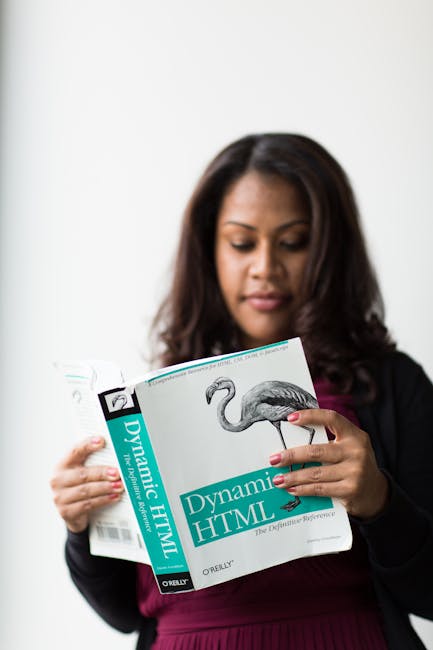 A woman with dark brown hair, wearing a black blazer over a burgundy top, is reading a book titled 'Dynamic HTML' by O'Reilly, which features an illustration of a flamingo on the cover. She is holding