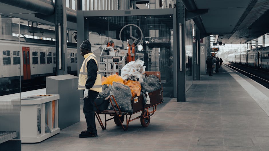 A person wearing a grey beanie, black jacket, and white gloves stands next to a large cart loaded with black, grey, and orange garbage bags and other waste materials on the platform of Welling train s